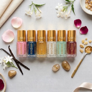 May include: A collection of six small, colorful perfume bottles with gold caps, arranged in a row. Each bottle has a different colored liquid and a decorative gold pattern. The image also includes flowers, petals, and vanilla beans.
