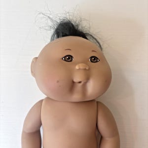 May include: A doll with a round, tan-colored vinyl head and body, featuring dark brown eyes, a small nose, and a closed mouth. The doll has sparse, dark black hair tied in a small tuft at the top of the head and a small mole on the left cheek.