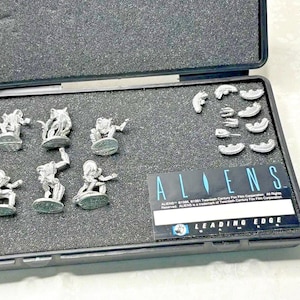 May include: A black case containing several silver figurines and small pieces. The figurines depict characters from the movie "ALIENS". The case also includes a card with the "ALIENS" logo and "LEADING EDGE" text.