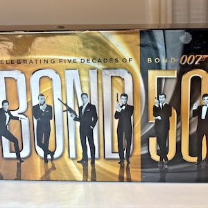 May include: A box set celebrating five decades of Bond 007. The design features the word "BOND" in large, silver letters, with the number "50" in gold. Silhouettes of James Bond actors are featured.