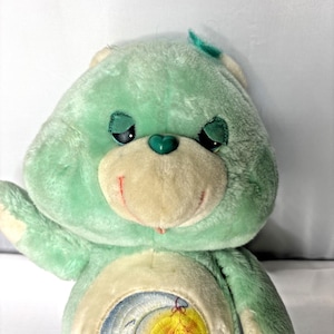 May include: A plush bear toy with mint green fur, a cream-colored muzzle, and a small heart-shaped nose. The bear features embroidered closed eyes with dark green eyelids, a stitched smile, and a circular graphic on its belly depicting a moon.