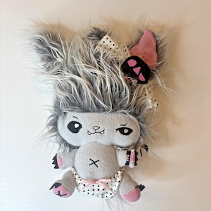 May include: A plush toy with long grey faux fur, a smooth grey fleece body, and pink accents on the ears and paws. It features black embroidered eyes, a small mouth with fangs, a white polka dot diaper with a pink ribbon, and a matching bow.