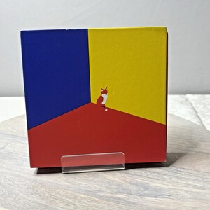 Puede incluir: Una caja cuadrada con un diseño minimalista que presenta una ilustración de un zorro. La caja está dividida en tres secciones: roja, amarilla y azul. El zorro está representado en un estilo simple y de dibujos animados, con vientre blanco y cola roja y blanca.