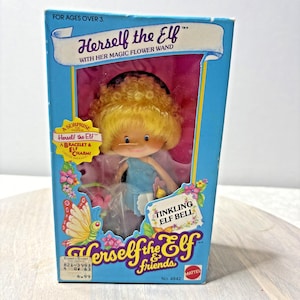Peut inclure: Une poupée vintage Herself the Elf dans sa boîte d'origine. La boîte bleue porte les mots "Herself the Elf" et "Tinkling Elf Bell". La poupée a des cheveux blonds et porte une robe bleue. La boîte comprend également une surprise "Bracelet & Elf Charms".