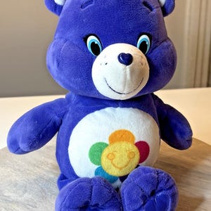 Puede incluir: Un peluche morado de Care Bear con una barriga blanca con un diseño floral colorido. El oso tiene un hocico blanco con una nariz en forma de corazón, ojos azules y detalles blancos en las orejas. Está sentado sobre una superficie de madera.