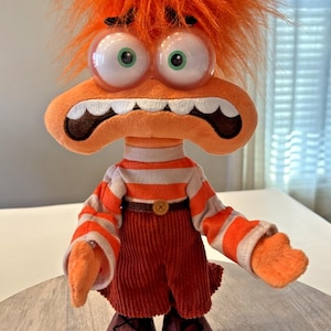 Op de afbeelding: Een pluche speelgoedfiguur met oranje haar, grote wiebelogen en een brede, grijnzende mond. Het draagt een gestreept shirt, corduroy broek en bruine schoenen. Het speelgoed heeft een verbaasde uitdrukking en lijkt op een stripfiguur.