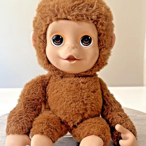 Op de afbeelding: Een pluche aap pop met een bruine vacht lichaam en hoofd. De pop heeft een licht getint gezicht met grote, expressieve ogen en een kleine, lachende mond. De armen en benen zijn ook licht getint.