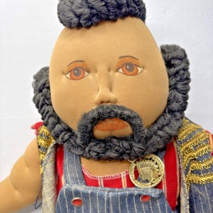 Puede incluir: Muñeca hecha a mano con rostro bronceado, ojos marrones y barba y cabello negros. La muñeca viste un atuendo a rayas rojas y azules con un medallón dorado. El atuendo incluye un peto vaquero azul con detalles rojos y dorados.