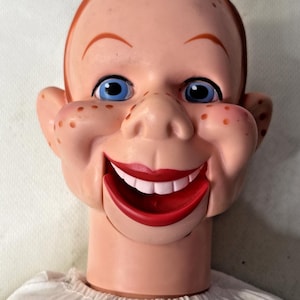 May include: A vintage ventriloquist dummy head with a smiling expression. The doll has blue eyes, red hair, and a red mouth. The face is flesh-toned with freckles. The doll is wearing a white shirt.