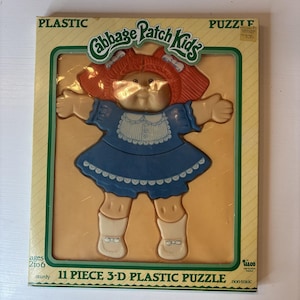 May include: An 11-piece 3-D plastic puzzle featuring a Cabbage Patch Kids doll. The doll has red hair, a blue dress, and white boots. The puzzle is in a yellow and green box with the words "Cabbage Patch Kids" and "Plastic" printed on it.