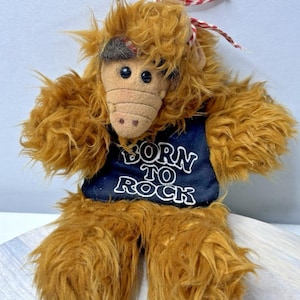 Op de afbeelding: Een pluche speelgoed van het personage ALF, met bruine vacht en een kenmerkend gezicht. Het draagt een marineblauw vest met de witte tekst "BORN TO ROCK". Een rood-wit gestreept lint is op zijn hoofd gebonden.