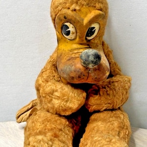 Op de afbeelding: Vintage pluchen hondenspeelgoed met een bruine, getextureerde body en een gele snuit. De hond heeft grote ogen, een zwarte neus en slappe oren. Het speelgoed zit rechtop, met de poten tegen elkaar. Een klassiek verzamelobject.