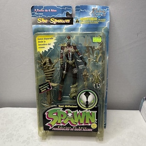 Op de afbeelding: She-Spawn actiefiguur in een doorzichtige plastic verpakking. De figuur is een vrouwelijk personage met een rood en wit masker, zwarte en bruine bepantsering en gouden accenten. De verpakking bevat de tekst "Todd McFarlane's Spawn" en "Edicion de Lujo Personajes de Gran Accion."