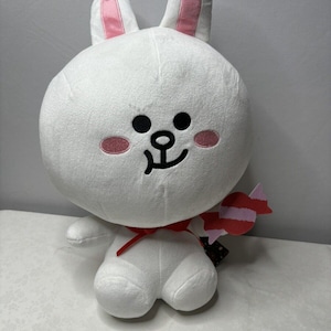 May include: A white plush bunny with pink inner ears, rosy cheeks, and a black smiling face. The bunny has a red bow and holds a candy-shaped accessory. The toy is designed with a cartoonish aesthetic.