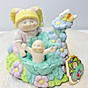 Puede incluir: Una figurita de porcelana de una muñeca Cabbage Patch Kid con coletas rubias y un vestido rosa, sosteniendo una muñeca Cabbage Patch Kid bebé. Las muñecas están sentadas en una hoja de col verde con flores y una mariposa.