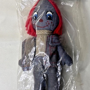 Op de afbeelding: Een vintage Joey Denim pop in een plastic zak. De pop heeft een denim lichaam, rood haar en een sjaal. De verpakking toont een stripfiguur op een motor. De tekst "Joey Denim" staat op de verpakking.