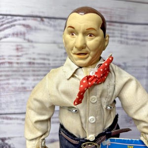 May include: A collectible doll of Curly Howard from The Three Stooges, featuring a moulded head with brown hair, a beige button-down shirt, a red neckerchief with white polka dots, and dark blue trousers with a brown belt and a visible metal buckle.