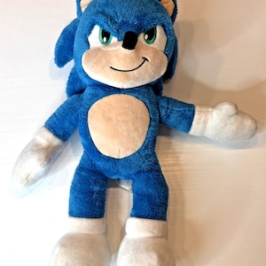 May include: A blue plush toy of Sonic the Hedgehog, featuring white hands, feet, and belly. The toy has green eyes, a black nose, and a smiling face. The toy is approximately 30 cm tall.