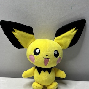 May include: A plush toy of a Pichu character, featuring a yellow body, large black-tipped ears, and pink cheeks. The toy has a black collar and a smiling face with a pink tongue. It is a soft, cuddly toy.