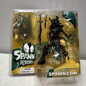 Op de afbeelding: Een Spawn Reborn actiefiguur van Domina, verpakt in een doorzichtige plastic hoes. De figuur is een krijger met zwarte bepantsering, turquoise accenten en een zwaard. De verpakking bevat het Spawn-logo en de website spawn.com.