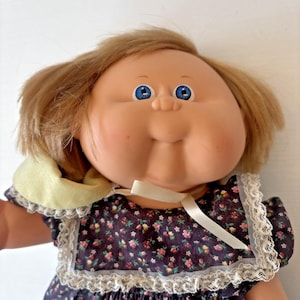 May include: A vintage doll with blonde hair and blue eyes. The doll has a round face with rosy cheeks and a slight smile. It is wearing a dark blue dress with a floral pattern and white lace trim. A white ribbon is tied around the neck.