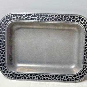 Puede incluir: Bandeja rectangular de metal con borde decorativo. La bandeja tiene un acabado plateado con un patrón negro tipo mosaico en los bordes. La bandeja está vacía y se encuentra sobre una superficie blanca.