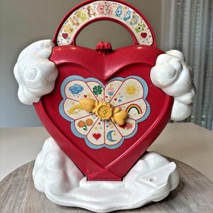 May include: A red heart-shaped toy with a handle featuring Care Bears. The toy has a white cloud base and cloud accents. The front has a flower design with a yellow bow and various symbols, including a rainbow, sun, and moon.