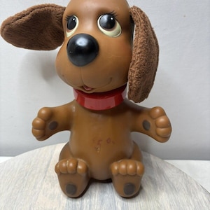 Puede incluir: Un perro de juguete de goma marrón con orejas caídas, ojos grandes y un collar rojo. El perro está sentado con las patas extendidas.