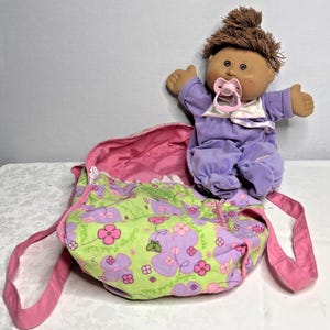 May include: A Cabbage Patch Kids doll wearing a purple outfit and a pacifier, with brown hair. The doll is next to a green and pink floral print diaper bag with pink straps.