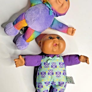 Puede incluir: Dos muñecas Cabbage Patch Kids. Una muñeca lleva un mono morado con detalles de arcoíris y una capucha a juego. La otra muñeca lleva un conjunto morado con un mono azul claro con estampado de búhos.