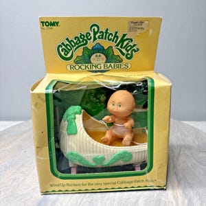 May include: A vintage Cabbage Patch Kids Rocking Babies toy in its original box. The box is yellow with green accents and features the Cabbage Patch Kids logo. The toy inside is a small baby doll in a white and green rocking cradle. The box reads "Wind-Up Rockers".