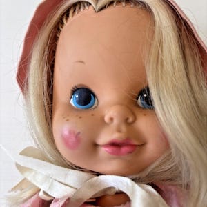 Puede incluir: Una muñeca vintage con cabello rubio y ojos azules. La muñeca tiene mejillas rosadas, pecas y lleva un vestido y un gorro rosas. Una cinta blanca está atada alrededor del cuello. La expresión de la muñeca es suave y acogedora.