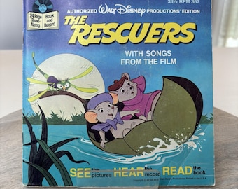May include: A vintage record book and record set featuring the Disney movie The Rescuers. The cover features the two main characters, Bernard and Miss Bianca, in a lily pad boat. The text on the cover reads "The Rescuers" with the tagline "With Songs From The Film".