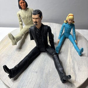 Op de afbeelding: Drie vintage actiefiguren zijn gerangschikt op een lichtgekleurd houten oppervlak. De figuren omvatten een inheemse Amerikaan in crèmekleurige kleding, een man in een zwart pak en een meisje in een blauwe overall. De figuren zitten.