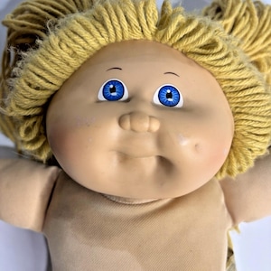 Könnte beinhalten: Eine Vintage-Puppe mit einem weichen, gebräunten Gesicht, rosigen Wangen und leuchtend blauen Augen. Die Puppe hat einen neutralfarbenen Körper und hellgelbes Garnhaar. Der Ausdruck der Puppe ist sanft und einladend.