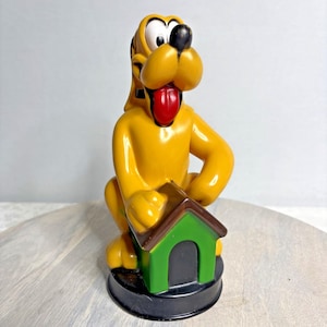 Könnte beinhalten: Eine gelbe Pluto-Figur, die ein grünes und braunes Hundehäuschen hält. Die Figur hat eine rote Zunge, schwarze Ohren und eine schwarze Nase. Der Sockel ist schwarz.