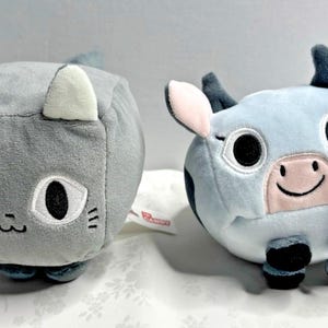 May include: Two plush toys: a gray cat with white ears and a blue cow with black spots. The cat has large eyes and a stitched mouth. The cow has a pink nose and a smiling mouth. Both are cube-shaped.