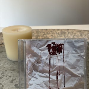Op de afbeelding: Een doorzichtig plastic CD-doosje met een hoesje met een gekreukelde zilveren achtergrond en een donkerrood, druipend inktontwerp in het midden bovenaan. De tekst "Full Disclosure" is in cursief schrift onderaan de hoes gedrukt.