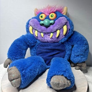 May include: A plush monster toy with blue fur, a purple face, and yellow teeth. The monster has green eyes, a green nose, and pink and purple hair. The toy is sitting on a light-colored surface.