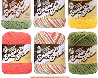 May include: Six balls of Sugar'n Cream yarn in various colors, including green, yellow, orange, and a striped pattern of red, white, green, and yellow. The yarn is labeled "The Original Sugar'n Cream" and is displayed with a wooden ruler and a crochet hook gauge.