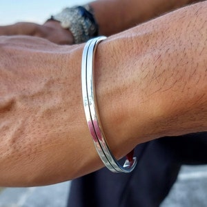 Puede incluir: Una pulsera plateada con un diseño de doble banda. La pulsera se lleva en la muñeca de una persona. La pulsera parece estar hecha de metal y tiene un acabado liso y pulido. La pulsera es una joya sencilla pero elegante. El fondo es el cielo.