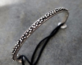 May include: A silver-colored bangle bracelet with a dotted pattern along the top edge. The bracelet is thin and curved, with a black string attached. The background is a textured gray surface.