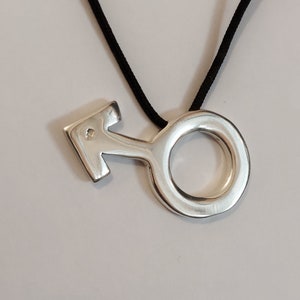 Puede incluir: Un collar con un colgante de plata en forma de s&iacute;mbolo masculino. El colgante tiene un peque&ntilde;o diamante en el centro.
