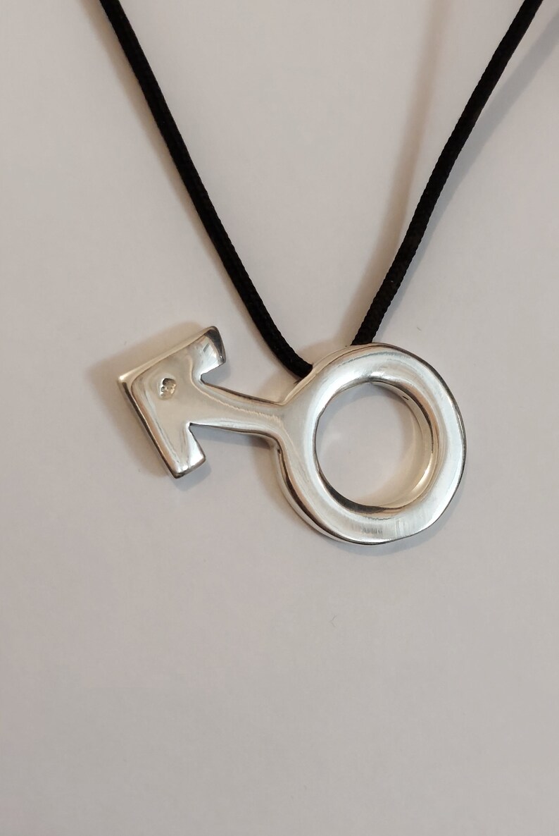 Puede incluir: Un collar con un colgante de plata en forma de s&iacute;mbolo masculino. El colgante tiene un peque&ntilde;o diamante en el centro.