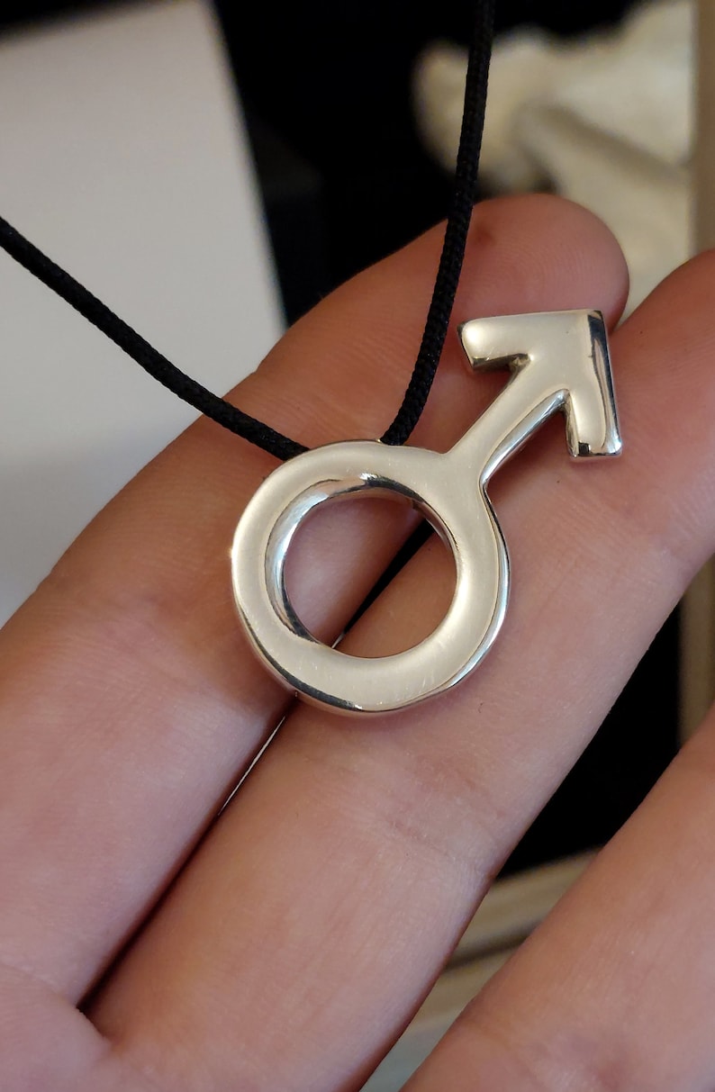 May include: A silver pendant necklace with a male symbol charm. The pendant is on a black cord.