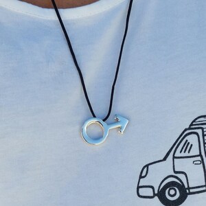 Puede incluir: Un collar de plata con un colgante de s&iacute;mbolo masculino en un cord&oacute;n negro. El collar lo lleva una persona que lleva una camiseta blanca con una ilustraci&oacute;n en blanco y negro de un coche y el texto "GREEN ROOM".