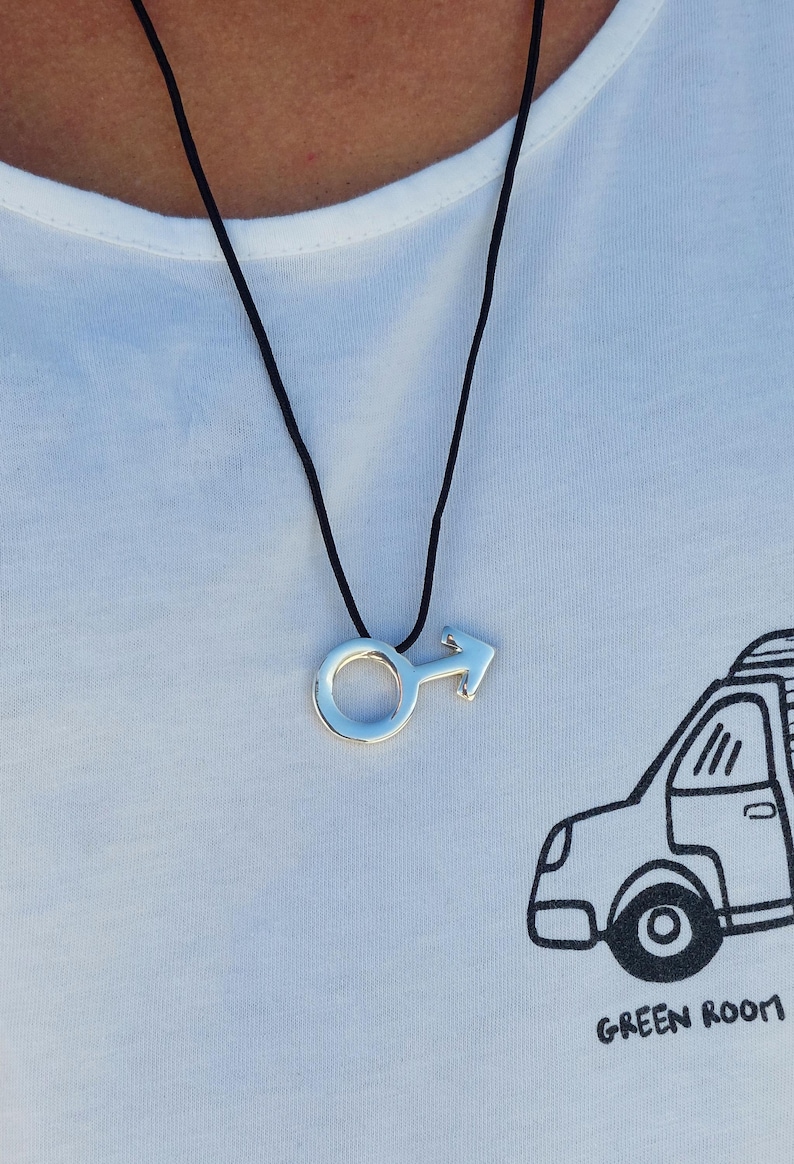 Puede incluir: Un collar de plata con un colgante de s&iacute;mbolo masculino en un cord&oacute;n negro. El collar lo lleva una persona que lleva una camiseta blanca con una ilustraci&oacute;n en blanco y negro de un coche y el texto "GREEN ROOM".