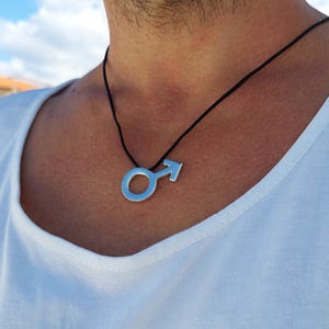 Puede incluir: Un collar de plata con un colgante de s&iacute;mbolo masculino en un cord&oacute;n negro.
