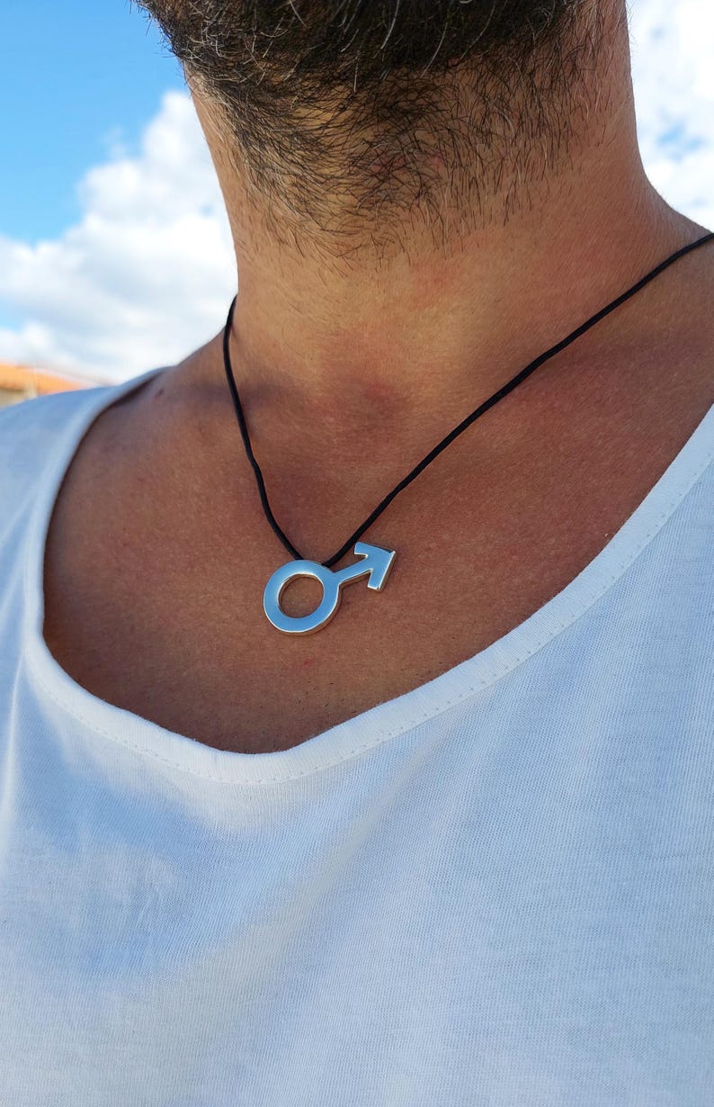 Puede incluir: Un collar de plata con un colgante de s&iacute;mbolo masculino en un cord&oacute;n negro.