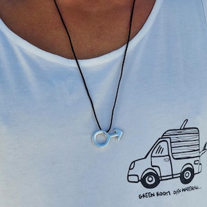 Puede incluir: Un collar de cord&oacute;n negro con un colgante plateado en forma de s&iacute;mbolo masculino. El collar lo lleva una persona que lleva una camiseta blanca de tirantes con una ilustraci&oacute;n en blanco y negro de una furgoneta con el texto "GREEN ROOM open waterfall..." en la parte delantera.